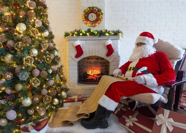 クリスマスツリー クリスマスストッキングや大きなギフトボックスの近くの彼の居心地の良い家の部屋でアームチェアに座っている古い種類の髭サンタクロースと子供のリストに書いて休日の準備 ロイヤリティフリーのストック画像