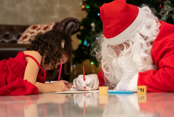 Santa Claus write a list of gifts to children on paper and little kid girl write a letter to Santa while lying together on floor in living room with Christmas tree at home
