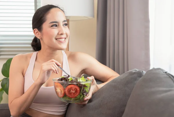 年轻的漂亮女人穿着运动服 享受丰盛的晚餐 在沙发上吃蔬菜沙拉 然后在家里快乐地做瑜伽锻炼 健康的生活方式和运动概念 — 图库照片#