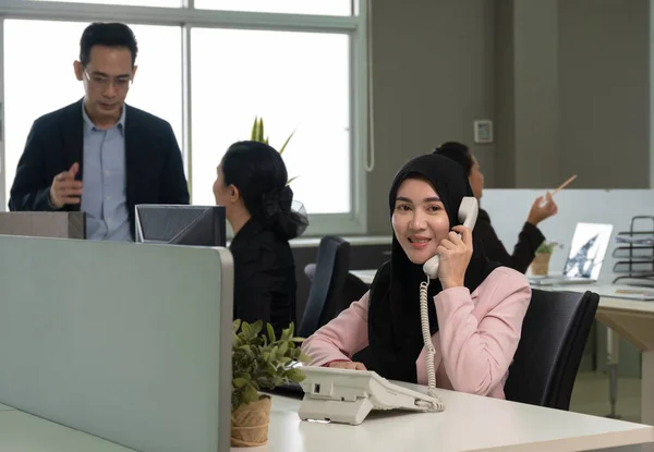 Muslim woman officer wearing hijab talking on landline phone while working on computer in busy open plan office. Colleagues busy working at desks in background