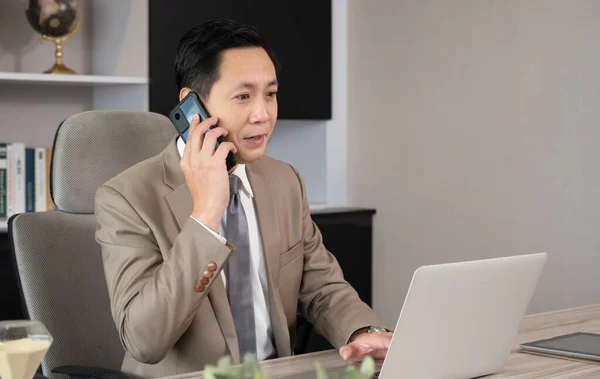 Company manager in brown suit having conversation on mobile phone. Businessman working on financial development strategy with laptop computer while sitting on desk in modern private office