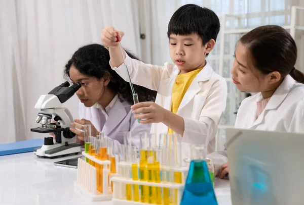 Pupil wearing white laboratory coat and learning science experiment by using eye dropper for dropping blue liquid to test tube in science laboratory room with scientist teacher. Children education concept with experiment, fun and enjoy class.