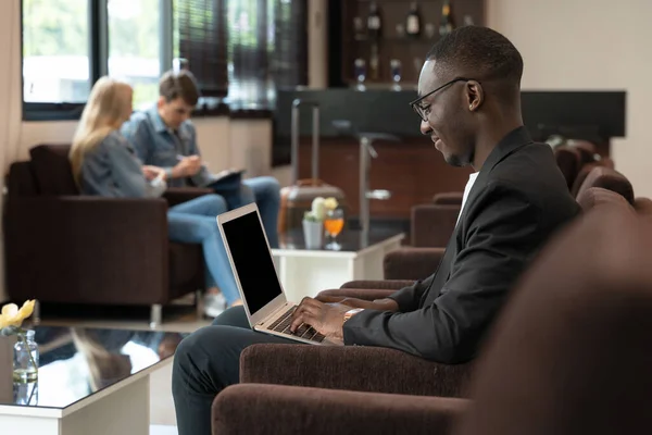African businessman sitting on sofa and waiting for the flight at airport lounge. Smiling man using PC laptop for presentation or new ideas of business project. Business and travel concept