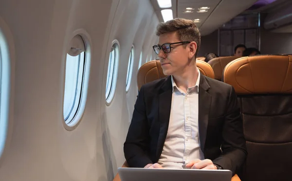 stock image Businessman in formal wear and glasses looking out of aircraft window during flight  while working with laptop computer. Internet connection on board service and business travel concept