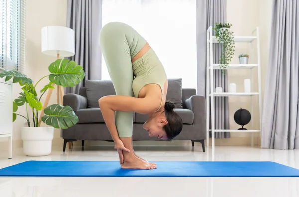Young female wearing green sportswear and practicing morning yoga routine at home, standing in forward fold with palms at the backs of ankles or Uttanasana to stretches spine, legs and hamstrings on blue mat in bedroom. Healthy life and fitness train