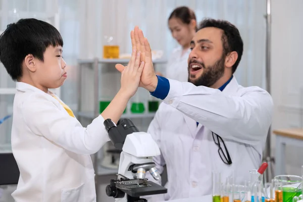Kid boy and scientist teacher wearing white laboratory coat and giving high five with raise hand up in science laboratory room. Children education concept with experiment, fun and enjoy class.