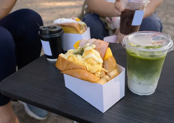 Breakfast toast sandwich with bacon, egg and cheese in take away box and cold drinks on wooden table at the park