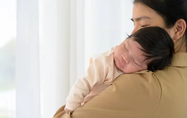 Adorable newborn baby girl sleeping on mom shoulder next to white curtain window. Young Asian mother hold infant 1 month with love, gently and happy. Safe with mom hug and healthy newborn baby concept