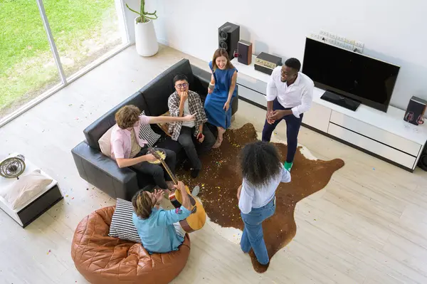 Group of young multiracial people having fun at house party, chatting with each other, playing acoustic guitar, singing, dancing and enjoy drinking in living room. Top view image. Youth lifestyle concept