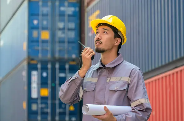 Japanese male engineer or foreman in uniform with yellow hardhat using walkie talkie to control loading containers at cargo shipping yard. Transportation and shipping concept