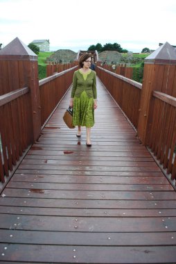Yeşil elbiseli bir kadın, kırsal manzarayla çevrili ahşap bir köprüde yürüyor. Dekor, mimari özellikler ve yeşilliğin bir karışımını sergiliyor ve sakin ve resimsel bir ortam yaratıyor..