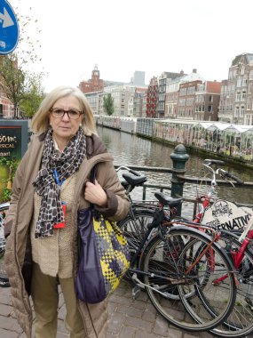 Kalın elbiseli bir kadın Amsterdam 'da bir kanalda duruyor. Arka planda bisikletleri var..