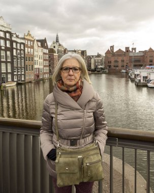 Hollanda, Amsterdam 'daki bir kanala bakan köprüde paltolu yaşlı bir kadın duruyor. Etrafı geleneksel binalarla çevrili..
