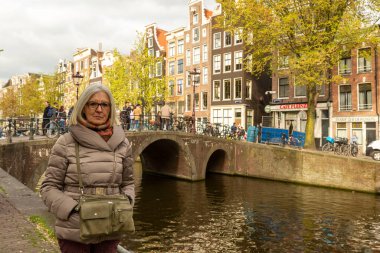 Hollanda, Amsterdam 'daki bir kanala bakan köprüde paltolu yaşlı bir kadın duruyor. Etrafı geleneksel binalarla çevrili..