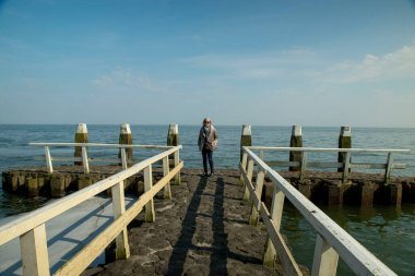 Hollanda 'da. Durgun mavi bir gökyüzünün altında, engin bir engin su alanına bakan bir iskelede duran biri. Sahne yalnızlığı, tefekküre dalmayı ve dışarıdaki güzelliği yansıtıyor..