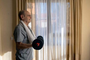 Üst düzey bir adam içeride bir dambıl kaldırıyor, dayanıklılık eğitimine ve refahına odaklanıyor. Güneş ışığı ve dingin ortam sağlıklı ve aktif bir yaşam tarzını vurgular, fitness veya motivasyon temaları için uygundur.