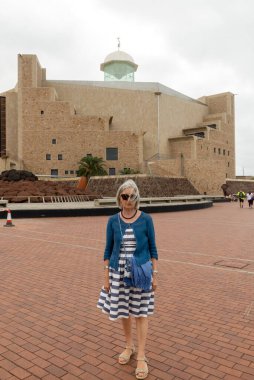 Alfredo Kraus Oditoryumu 'nun, Las Palmas de Gran Canaria' nın önünde bulutlu bir gökyüzünün altında, çağdaş mimari ve gündelik, şık giysiler sergileyen bir kadın duruyor..