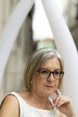 Gözlüklü ve kısa gri saçlı son sınıf öğrencisi bir kadın açık havada, derin düşüncelere dalmış oturuyor. Bilgelik ve içgözlem yayıyor samimi, kentsel bir ortamda, günlük yaz kıyafetleri giyiyor..