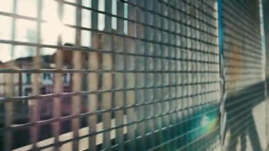 Kamera metal bir ızgaraya paralel hareket eder. Arka planda, odak dışı bazı binalar vardır.