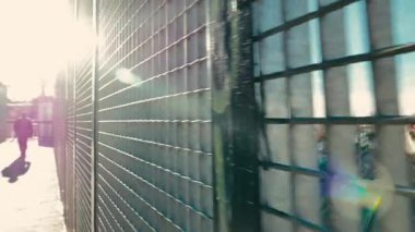 Odaklanamayan ve arkadan ışıklandırılmış bir adam kaldırımda yürüyor. Metalik tel örgüye odaklan. seyahat