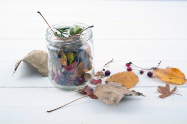 Kavanozda kurumuş yapraklar ve beyaz ahşap masada kırmızı böğürtlenler var.