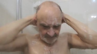 Yaşlı bir adam duş alırken yüzünü su ve sabunla yıkarken görülüyor. Görüntü, günlük bakım ve basitliği yansıtan rutin bir kişisel hijyen anını yakalıyor...