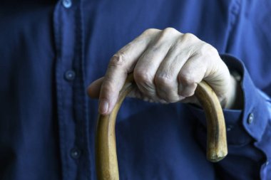 Yaşlı bir bireyin, tahta bastonu tutan, desteği, yaşlanmayı ve hareket kabiliyetini sembolize eden, bakım, yardım ve ihtiyarlık gibi kavramları temsil eden, yaşlı bir bireyin elinin yakın plan görüntüsü..