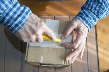 Sarı bir bıçakla karton bir kutunun mührünü açan kişi dikkatli bir şekilde tutuş ve ambalaj tasvir ediyor. Bu etkinlik, paketlerin açılması, teslimatı ve ilk süreci temsil eder..