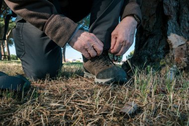 Kuru çimenlerin üzerinde diz çökerken patika çizmeleri bağlayan, doğa yürüyüşüne ya da maceraya hazırlanan birinin yakın çekimi.