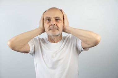 Olgun erkek, hafif bir arka plana karşı poz veriyor, kulaklarını kapatarak jestler yapıyor, hayal kırıklığını ya da huzursuzluğu ifade ediyor, dış ses veya dikkat dağınıklığından kaçınıyor, bu görüntüyü düşündürücü yapıyor..