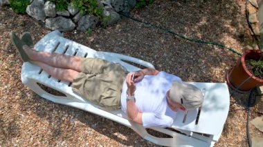 Bir birey rahat bir şekilde açık hava şezlongunda uzanır, sakin bir ortamın tadını çıkarırken çakıl ve yeşillik de dahil olmak üzere doğal elementlerle çevrili bir akıllı telefon kullanır..