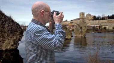 Zamora, İspanya. Arka planda tarihi mimari olan manzaralı bir nehir manzarasını fotoğraflamak için kamera kullanan bir adam, sakin ve kültürel bir ortam sergiliyor. Fotoğraf, seyahat ve tarihi keşifler için idealdir..