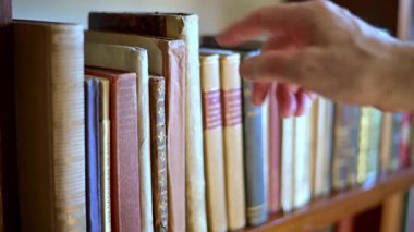 A gentle hand reaches for a weathered book on a wooden library shelf, capturing a moment of curiosity, learning, and nostalgia in a warm, quiet home library scene.