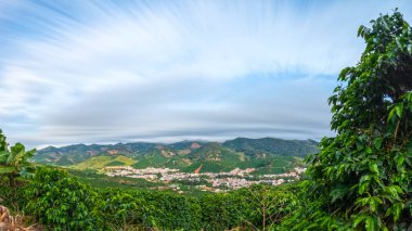 Alto Caparo 'nun dağlık yamaçlarda kahve ekinleri, yeşil bitki örtüsü ve vadide bir kasaba olan kırsal manzarası. Soyut bulutlarla uzun pozlama.