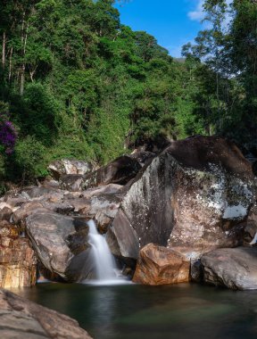 Bereketli yeşillikler ve tropikal yağmur ormanlarındaki devasa kayalarla çevrili tenha bir şelalenin huzurunun tadını çıkar. Zümrüt yeşili su serinlemek için mükemmel bir havuza rahatça akar.
