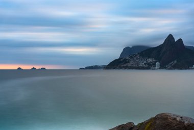 Rio de Janeiro, Arpoador Kayası 'nda çarpıcı bir günbatımı Dois Irmaos dağlarının gölgesine, sakin okyanusa ve çarpıcı mavi-turuncu gökyüzüne bakıyor..