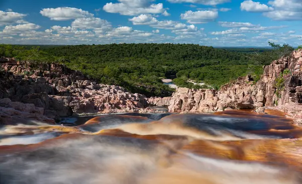 Aerial view of beautiful red waterfalls flowing into pools surrounded by vibrant greenery under a cloudy sky.