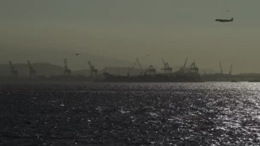 Uçak deniz kenarındaki havaalanına iniyor, endüstriyel vinçler arka planda.