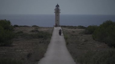 Bisiklet ve motosikletli turistler deniz kıyısındaki tenha bir deniz fenerine giden bir kıyı yolunda toplanıyorlar..