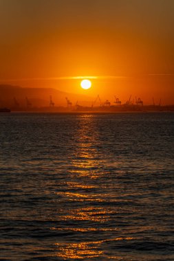 Gün batımında turuncu bir günbatımına karşı çizilmiş vinçleri ve gemileri olan sanayi limanı..