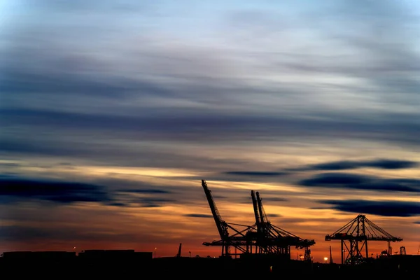 Commercial dock bustling with activity under a vibrant dusk sky, creating a contrast between the silhouettes of cranes, containers, and ships.