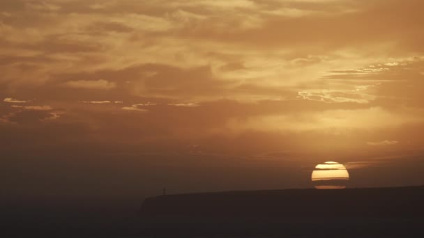 息をのむような夕日には 黄金の空に向かって崖と灯台のシルエットで雲の背後にある顕著な太陽の設定が特徴です — ストック動画