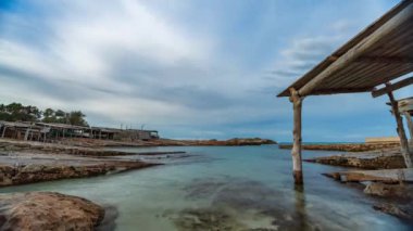 Formentera 'nın şeffaf sularında, mavi gökyüzünde hareket eden bulutlarla birlikte eski bir yarı dairesel iskelenin hızlandırılması..