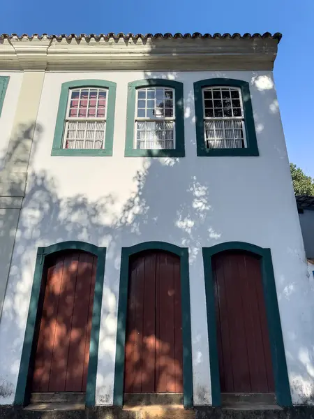 stock image Charming white house with green-trimmed windows and red doors, casting playful shadows.