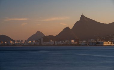 Rio de Janeiro Körfezi 'nin alacakaranlık fotoğrafında Kurtarıcı İsa' nın silueti, sahil evleri ve Dos Irmaos dağının uzun pozlardan oluşan ipeksi su etkisi görülüyor..