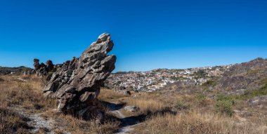 A towering rock formation stands before a tranquil hillside village beneath a clear blue sky. clipart