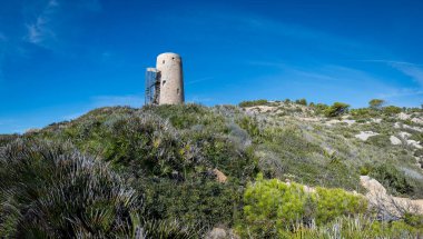 Eski bir taş gözetleme kulesi, berrak mavi gökyüzünün altındaki yemyeşil Akdeniz çalılarının arasında duruyor..