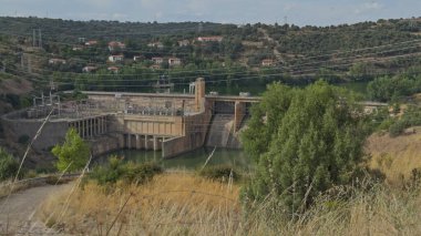 Derin bir kanyonda, hareket eden ağaç dalları ve doğal manzarayla çevrili bir hidroelektrik santralinin geniş görüntüsü..
