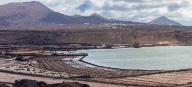 Kıyı tuz düzlükleri, bulutlu bir gökyüzü altında dağlar, uyumu ve endüstriyel karşıtlığı vurguluyor..