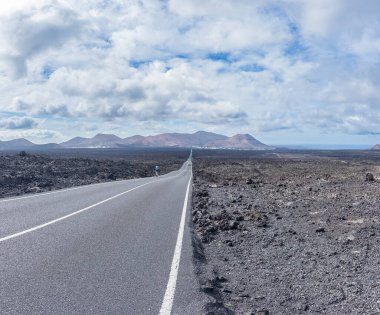 Issız bir volkanik araziyi tek başına geçen bir yol bulutlu bir gökyüzünün altındaki uzak dağlara doğru ilerliyor..